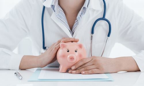 female doctor in white lap coat and stethoscope holding piggy bank over paperwork